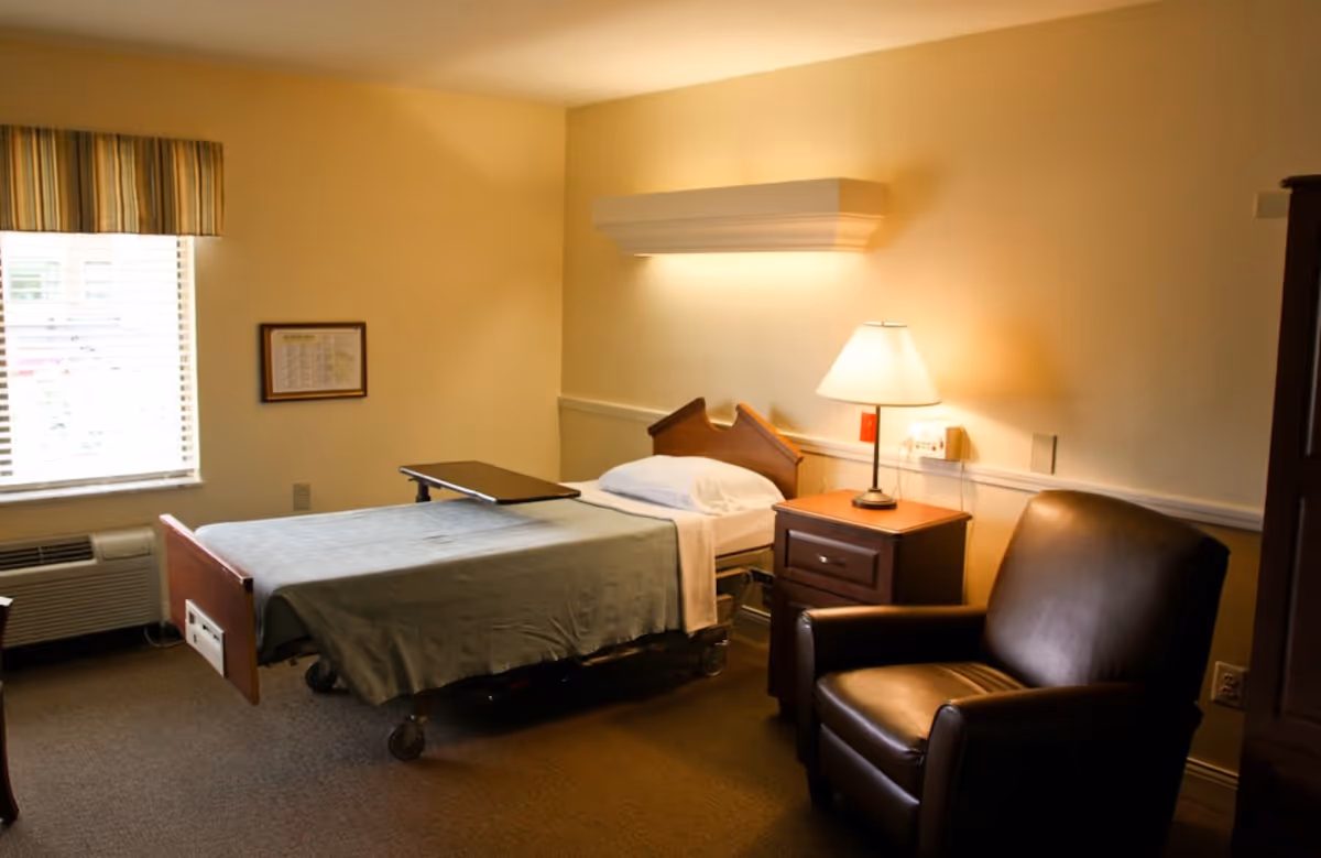 A small, neatly arranged bedroom with a single hospital-style bed covered with a gray blanket and a white pillow. Next to the bed is a wooden nightstand with a lamp on it, and beside the nightstand is a dark brown leather armchair. A window with striped curtains allows natural light into the room, and there is an air conditioning unit below the window. The walls are painted a light beige color.