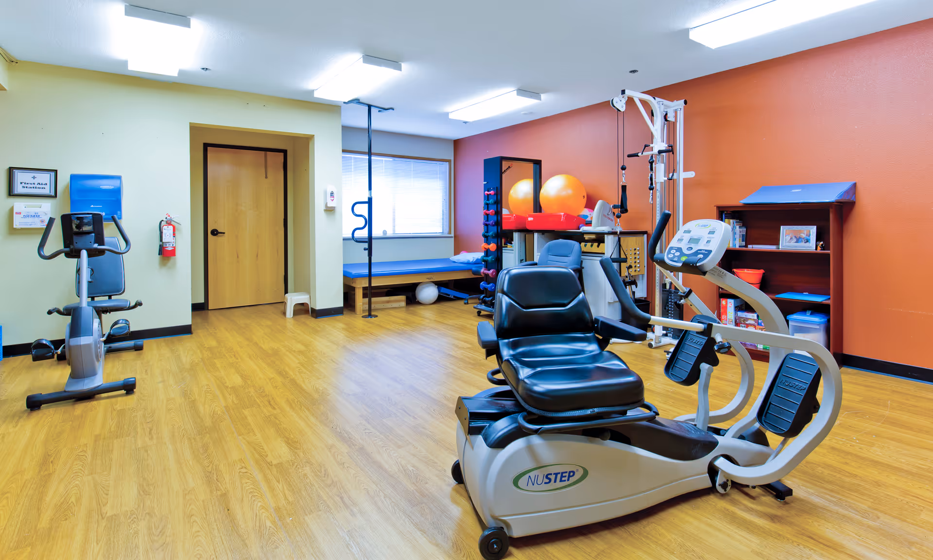 A bright indoor exercise/therapy room with a recumbent exercise machine, stationary bike, therapy table, and exercise balls.