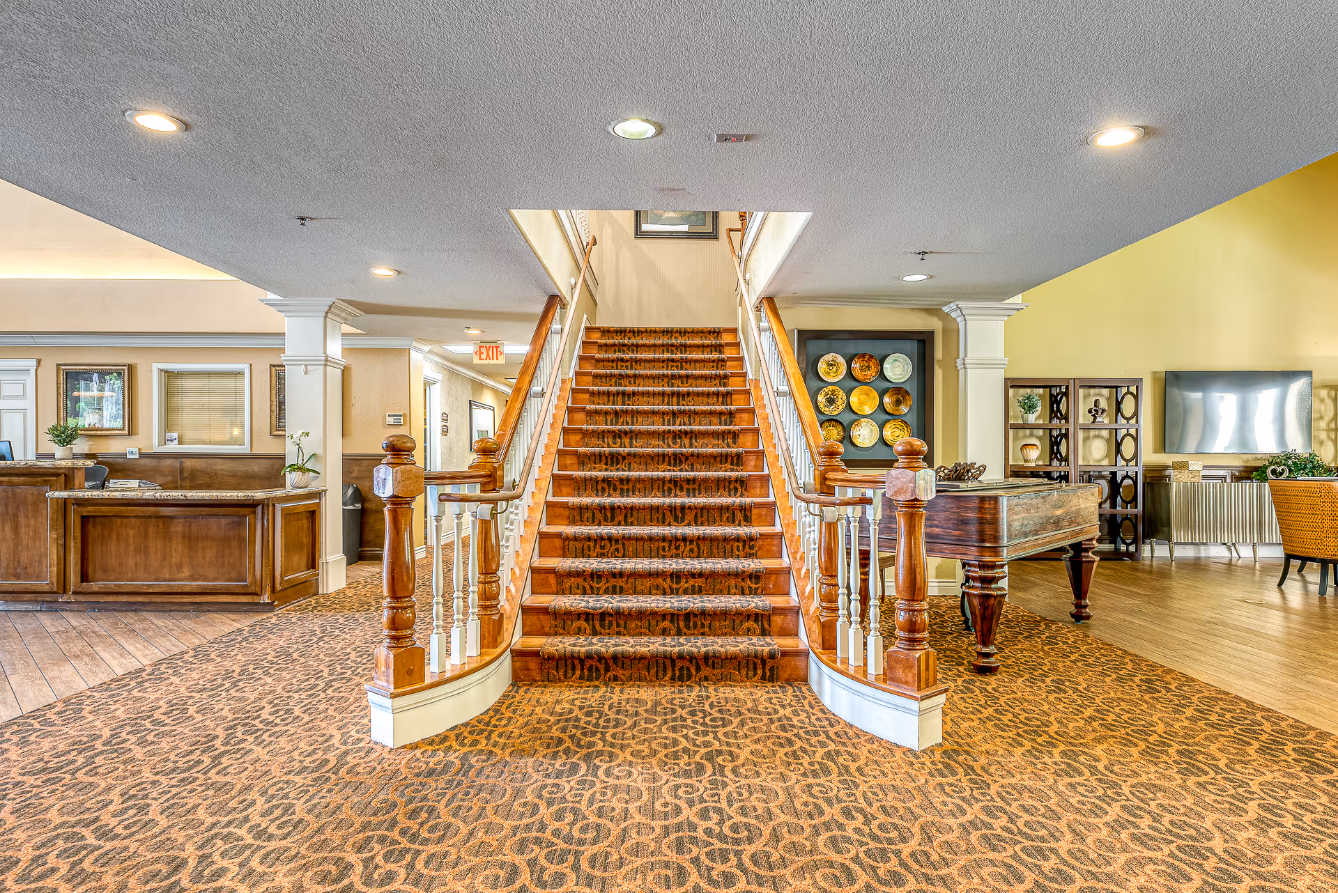 Interior view of a senior living facility lobby featuring a central carpeted staircase with wooden handrails, a reception desk on the left, decorative plates on the wall to the right, a wooden piano, and a seating area with a TV in the background.
