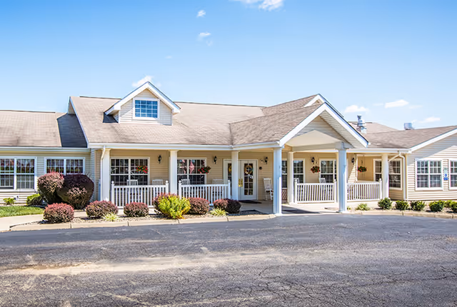 Front exterior view of a single-story building with beige siding, white trim, and a covered entrance supported by columns. There are several windows with flower boxes and neatly trimmed bushes along the front. The sky is clear and blue.