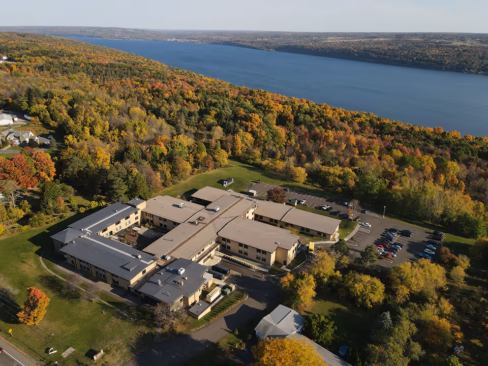 Aerial view of Cayuga Nursing & Rehabilitation Center surrounded by trees with autumn foliage, adjacent to a large body of water under a clear sky.