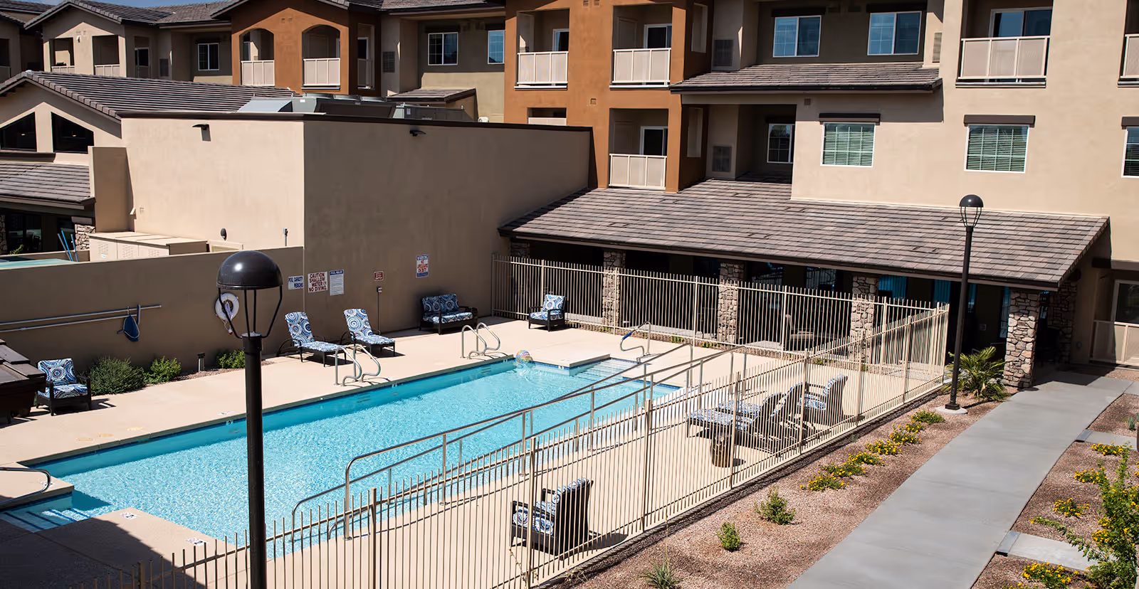 Outdoor swimming pool area at Westgate Village with lounge chairs and seating around the pool, enclosed by a metal fence. The pool is surrounded by a beige building with balconies and windows, and there are two lamp posts along a paved walkway with some landscaping.