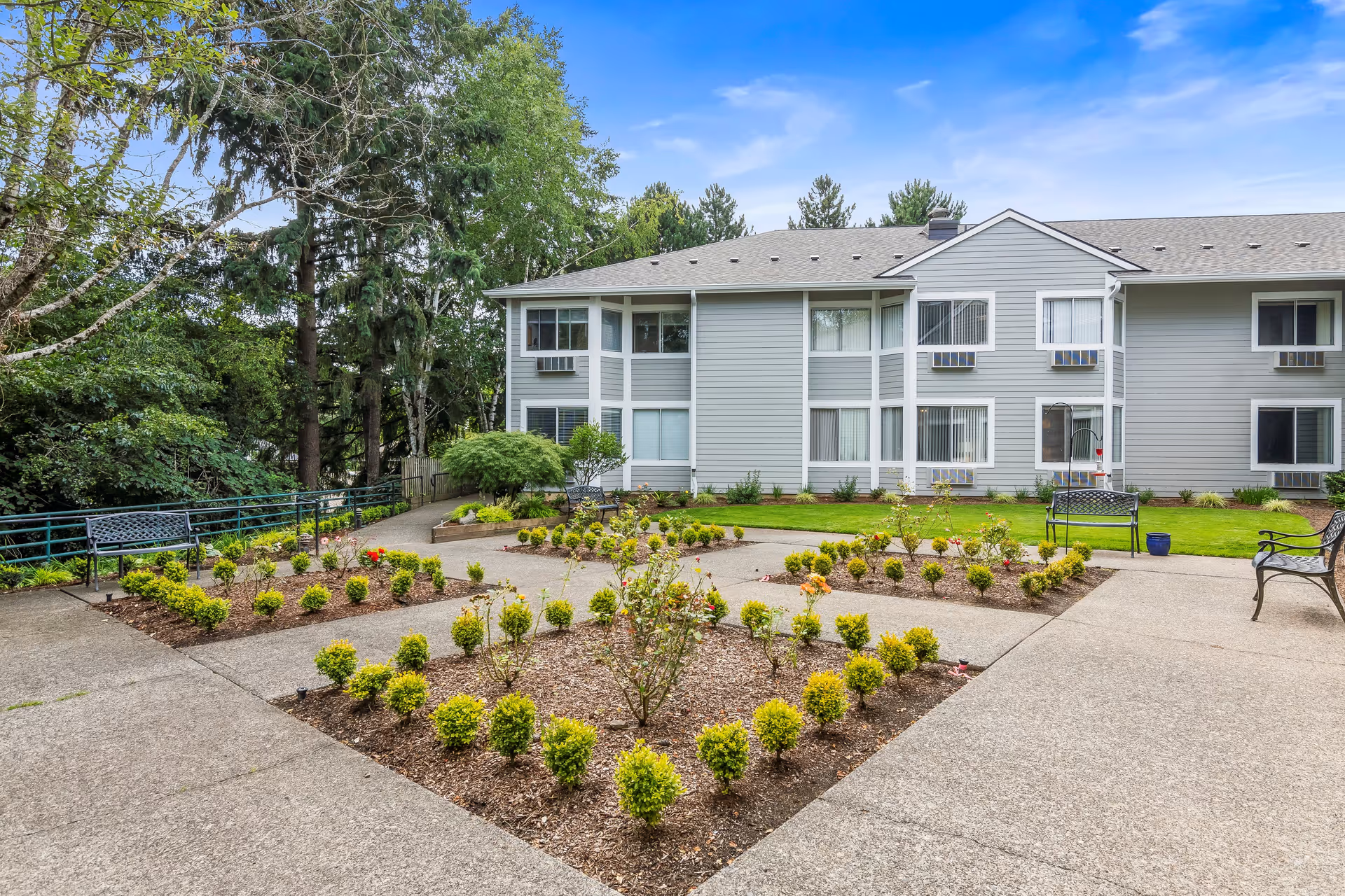 Outdoor courtyard area of a senior living facility with neatly trimmed bushes and flower beds arranged in geometric patterns, surrounded by paved walkways and benches. The background shows a two-story gray building with multiple windows and air conditioning units, set against a backdrop of tall green trees under a blue sky with some clouds.