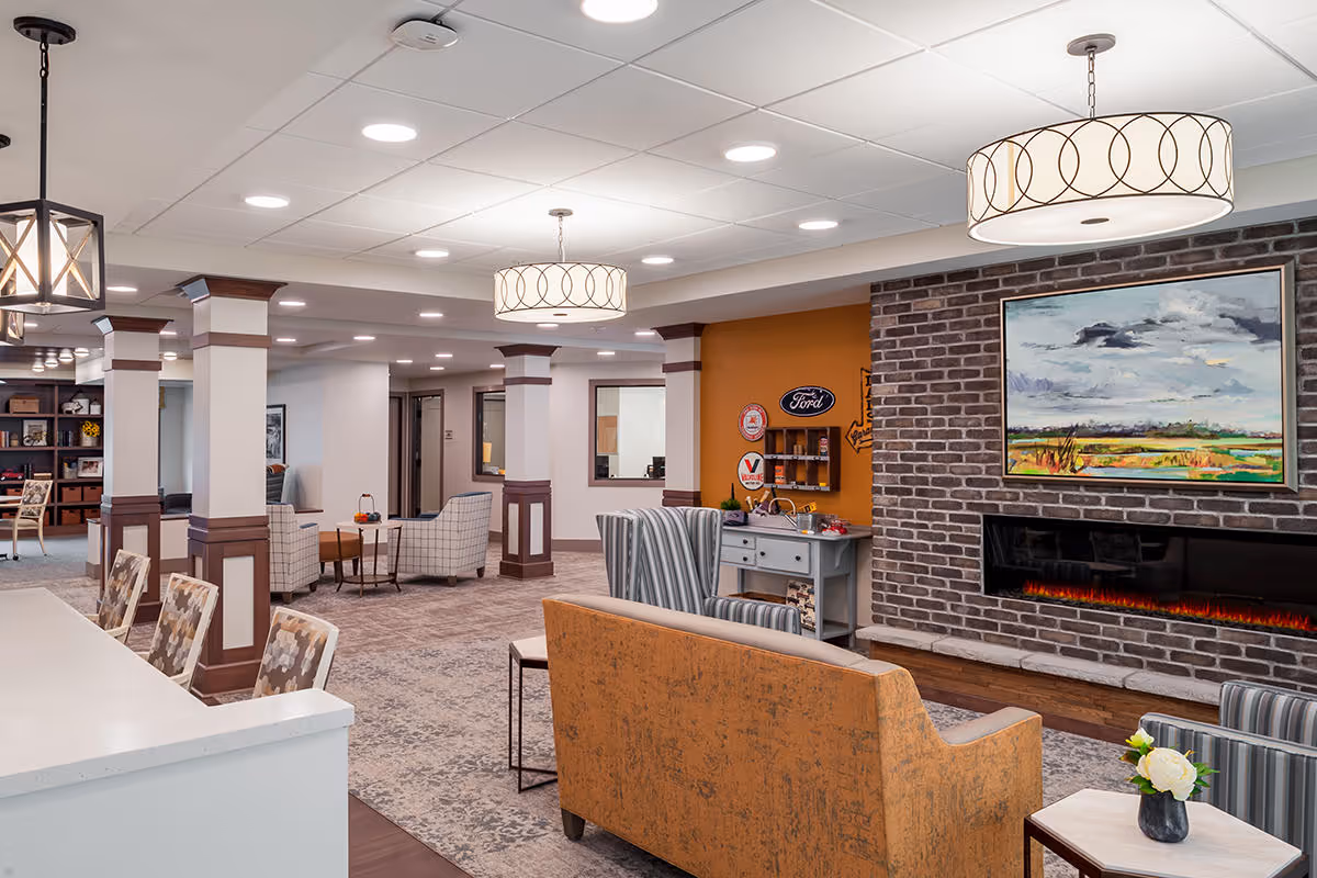 A spacious and well-lit living area in a senior living facility featuring comfortable seating including patterned armchairs and a mustard-colored sofa. The room has a modern electric fireplace set in a brick wall with a landscape painting above it. There are decorative light fixtures hanging from the ceiling and a small table with a flower vase in the foreground. The space includes columns with wood paneling and an open layout leading to other rooms.