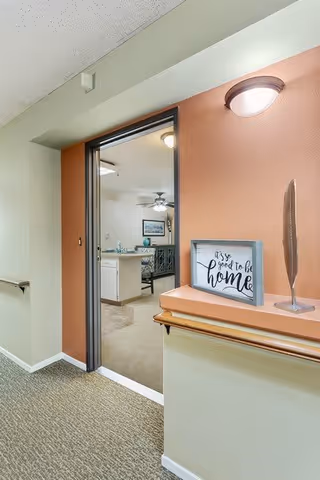 View of a hallway leading to an open doorway revealing a kitchen and dining area. The hallway has beige carpet and light green walls with a salmon-colored accent wall around the doorway. A small shelf on the accent wall holds a framed sign that reads 'It's so good to be home' and a decorative feather sculpture. A wall-mounted light fixture is above the shelf.