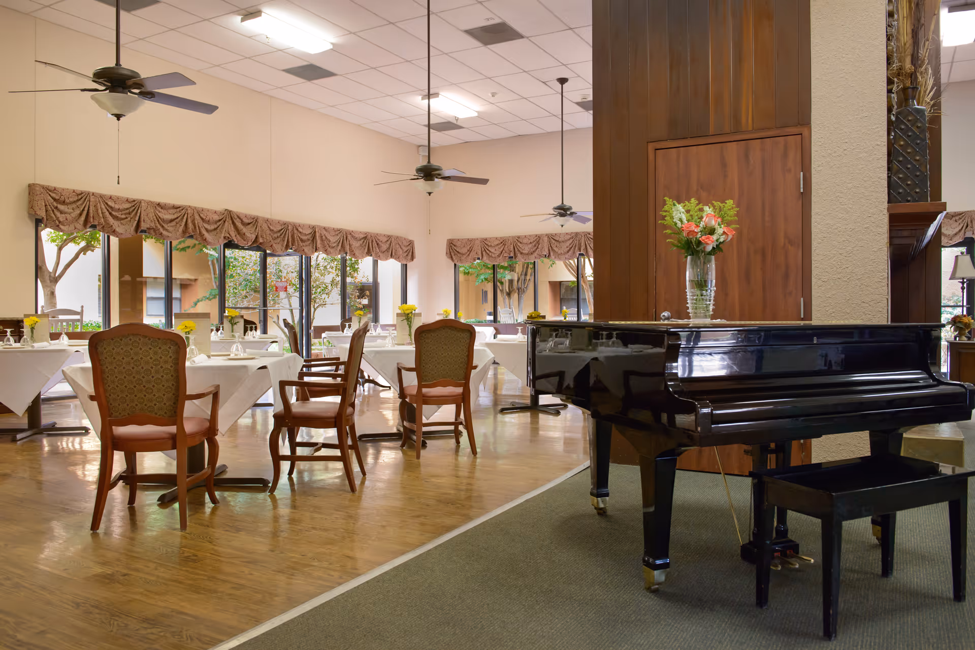 A dining room with several tables covered with white tablecloths and set with glasses and napkins. Wooden chairs with cushioned seats surround the tables. Large windows with valance curtains allow natural light to enter. A black grand piano with a vase of flowers on top is positioned on a carpeted area near a wooden wall with a closed door. Ceiling fans hang from the ceiling.