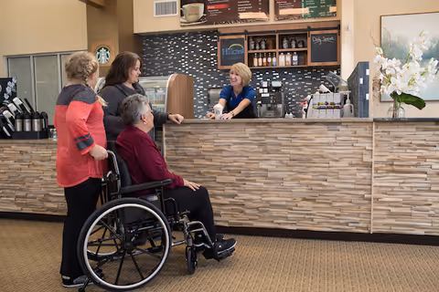 A woman in a wheelchair and another woman standing beside her are at a counter being served by a smiling female barista in a coffee shop area inside a senior living facility. The counter has a stone facade, and behind it are coffee machines, a menu board, and shelves with coffee supplies. A Starbucks logo is visible on the left side.