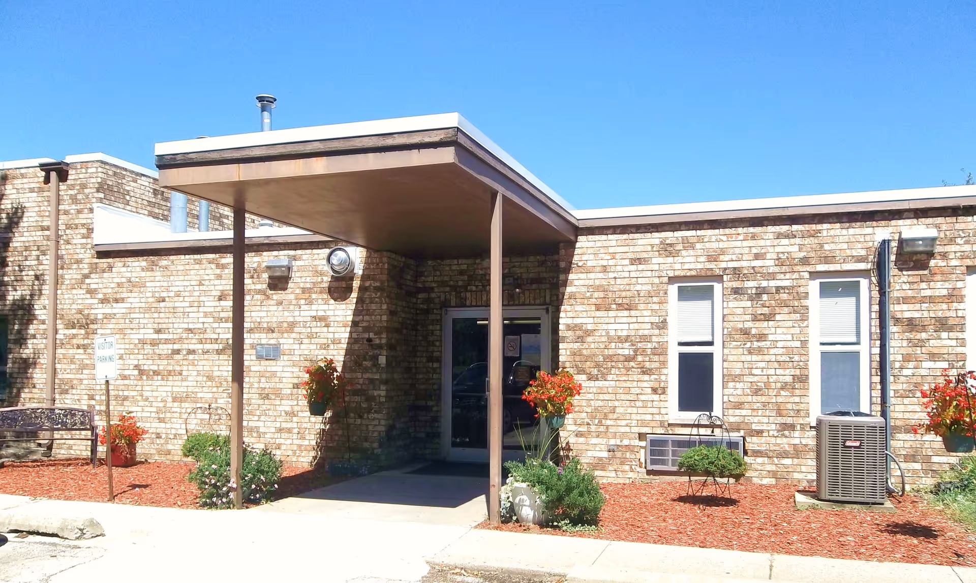 Exterior view of a single-story brick building with a covered entrance supported by two metal poles. There are two windows with white blinds, an air conditioning unit, and several flower pots with red flowers and green plants around the entrance. A sign indicating visitor parking is visible on the left side near a bench.