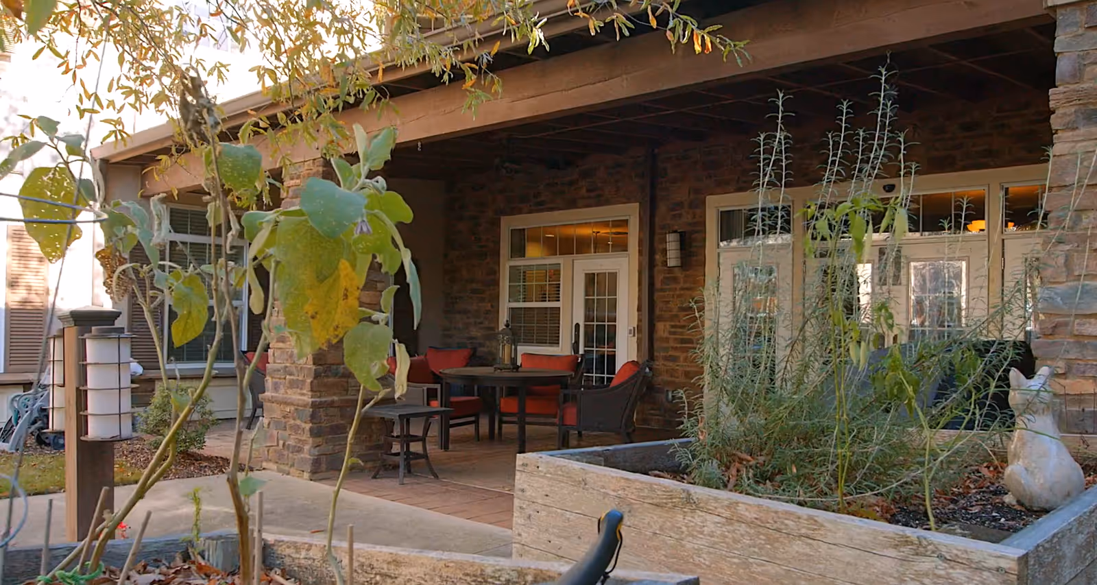 Outdoor covered patio area with a round table and four chairs with red cushions. The patio is attached to a building with stone walls and white-framed glass doors and windows. In the foreground, there are raised garden beds with plants and a small white cat statue.