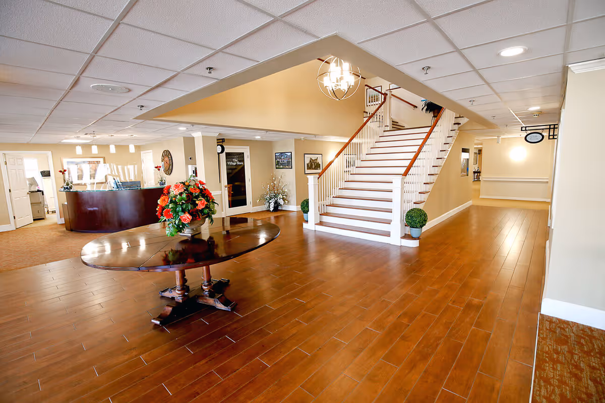 Spacious interior lobby area of The Village at Willow Crossings featuring a wooden round table with a floral arrangement in the center, a staircase with white railings and wooden handrails leading to the upper floor, a reception desk in the background, beige walls, and wood-look flooring.
