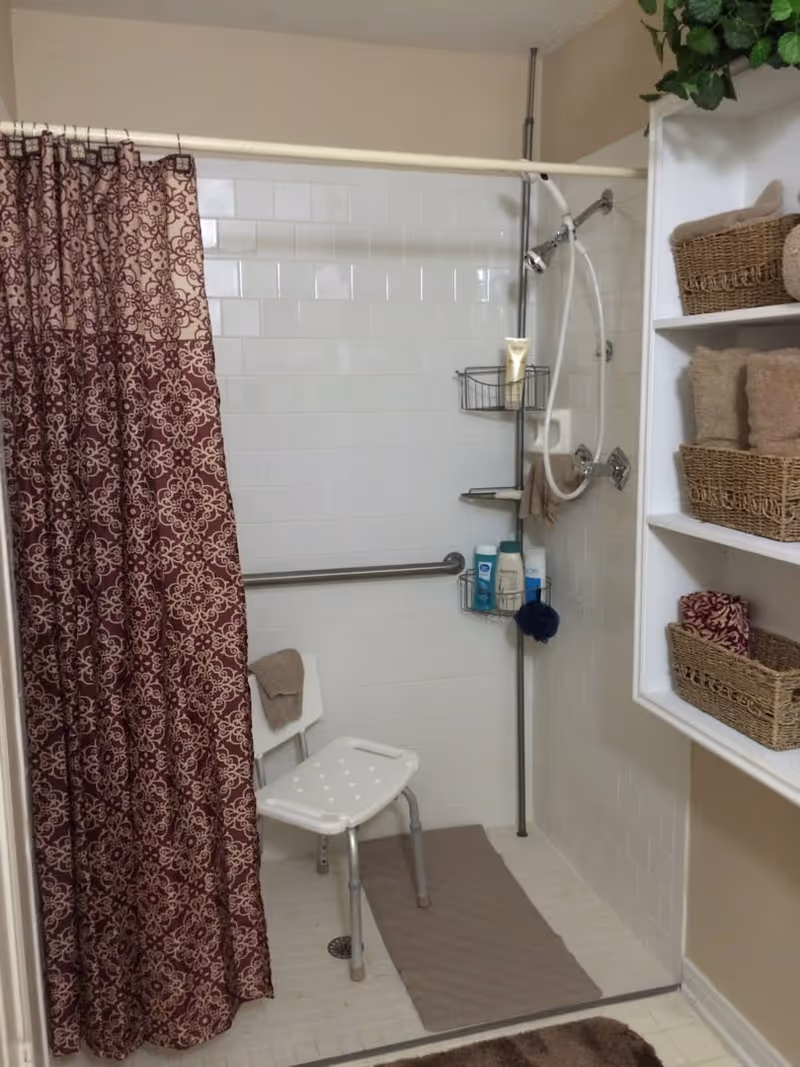 A bathroom shower area with white tiled walls and floor, featuring a maroon patterned shower curtain, a white shower chair, grab bars, a handheld showerhead, and shelves with toiletries and woven baskets holding towels.
