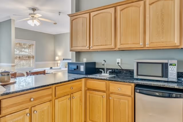 A kitchen area with wooden cabinets, a black granite countertop, a small sink, a microwave, and a mini dishwasher. In the background, there is a glimpse of a living or bedroom area with a ceiling fan, window with blinds, and a bed with a lamp on a nightstand.