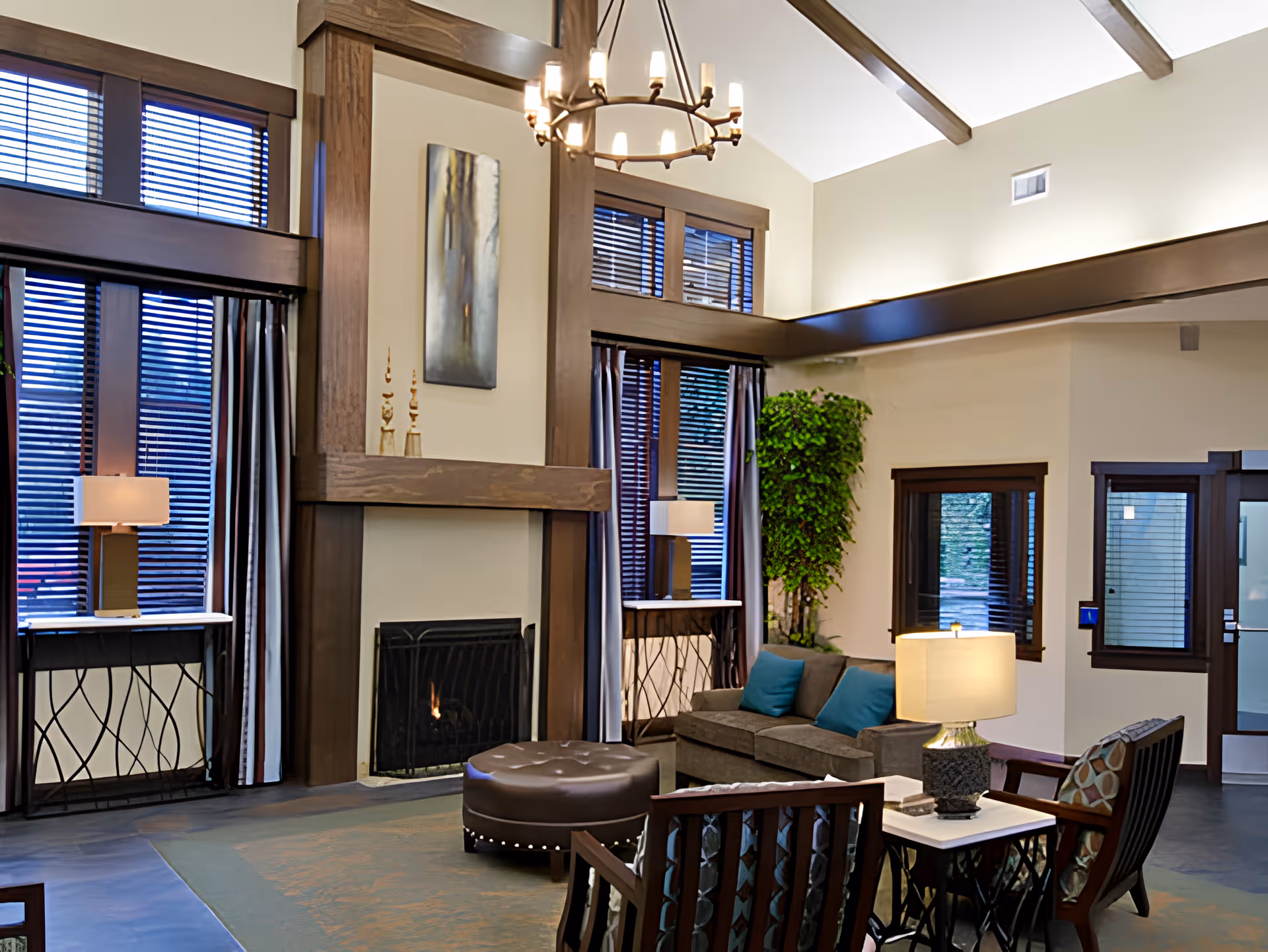 High-ceilinged lounge with a fireplace, sofa and chairs, lamps, and large windows with blinds.