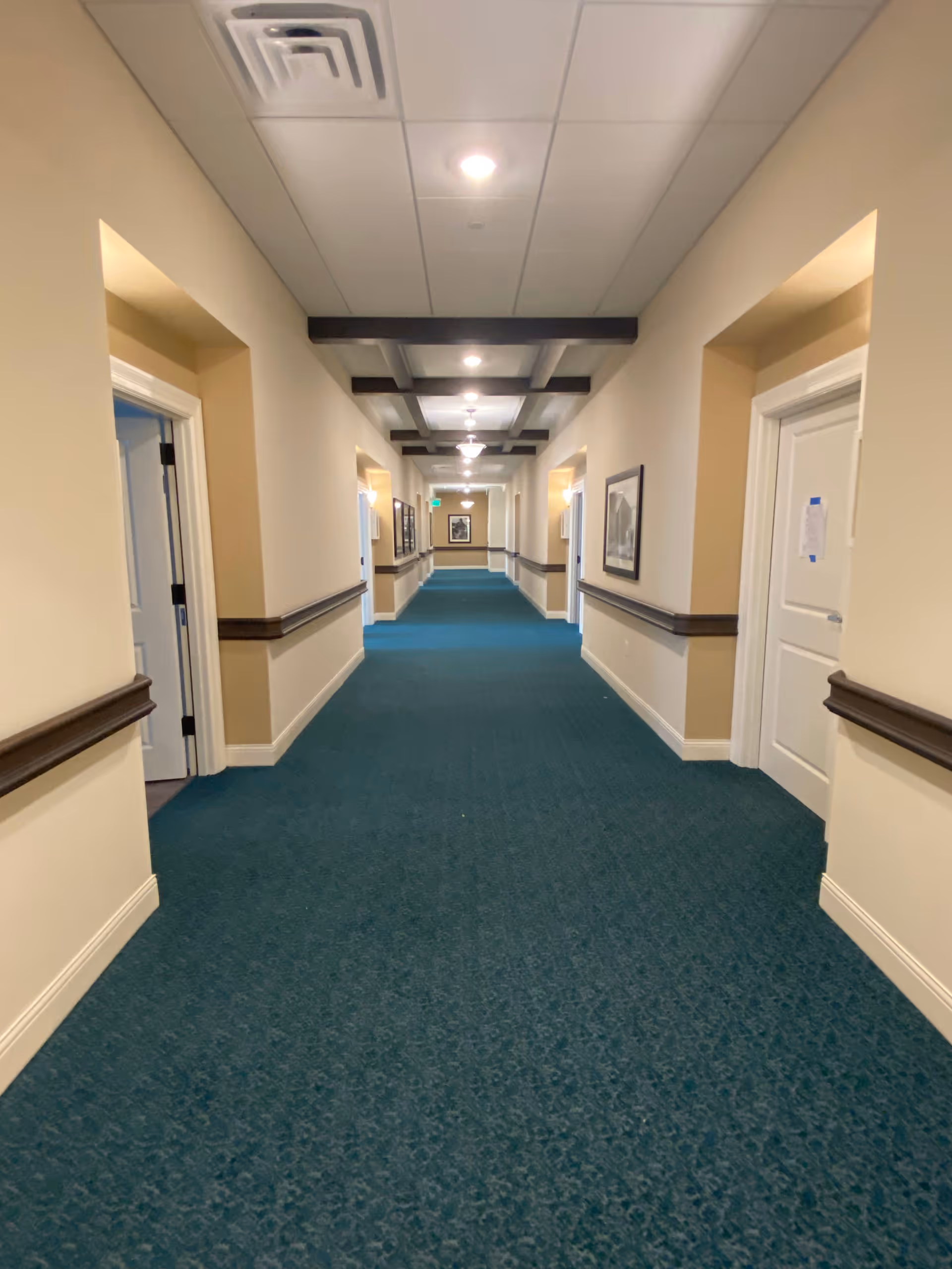 Long, carpeted interior hallway in a senior living facility with teal carpet, beige walls, overhead lighting, doors and framed pictures along the sides.