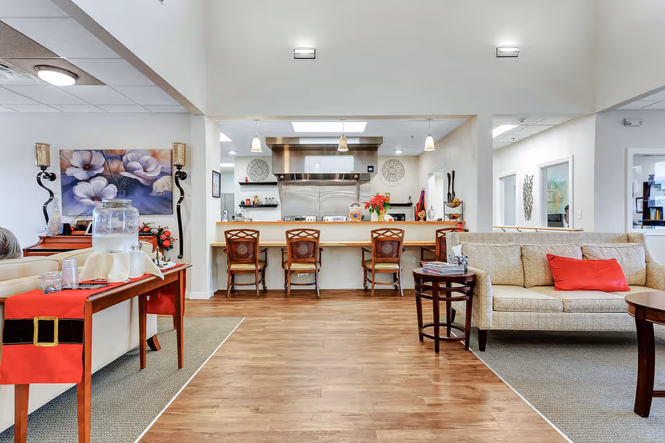 Interior view of a senior living facility showing a common area with a beige couch with a red pillow, a wooden table with chairs, and a kitchen counter with four wooden chairs. The kitchen area has stainless steel appliances and pendant lights hanging from the ceiling. There is a table with a red table runner and a water dispenser on the left side, and floral artwork on the wall.