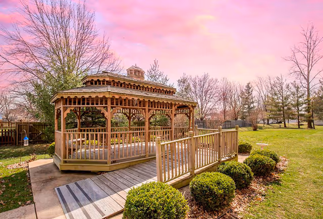A wooden gazebo with a ramp surrounded by green bushes and trees under a pink and purple sky in an outdoor garden area.