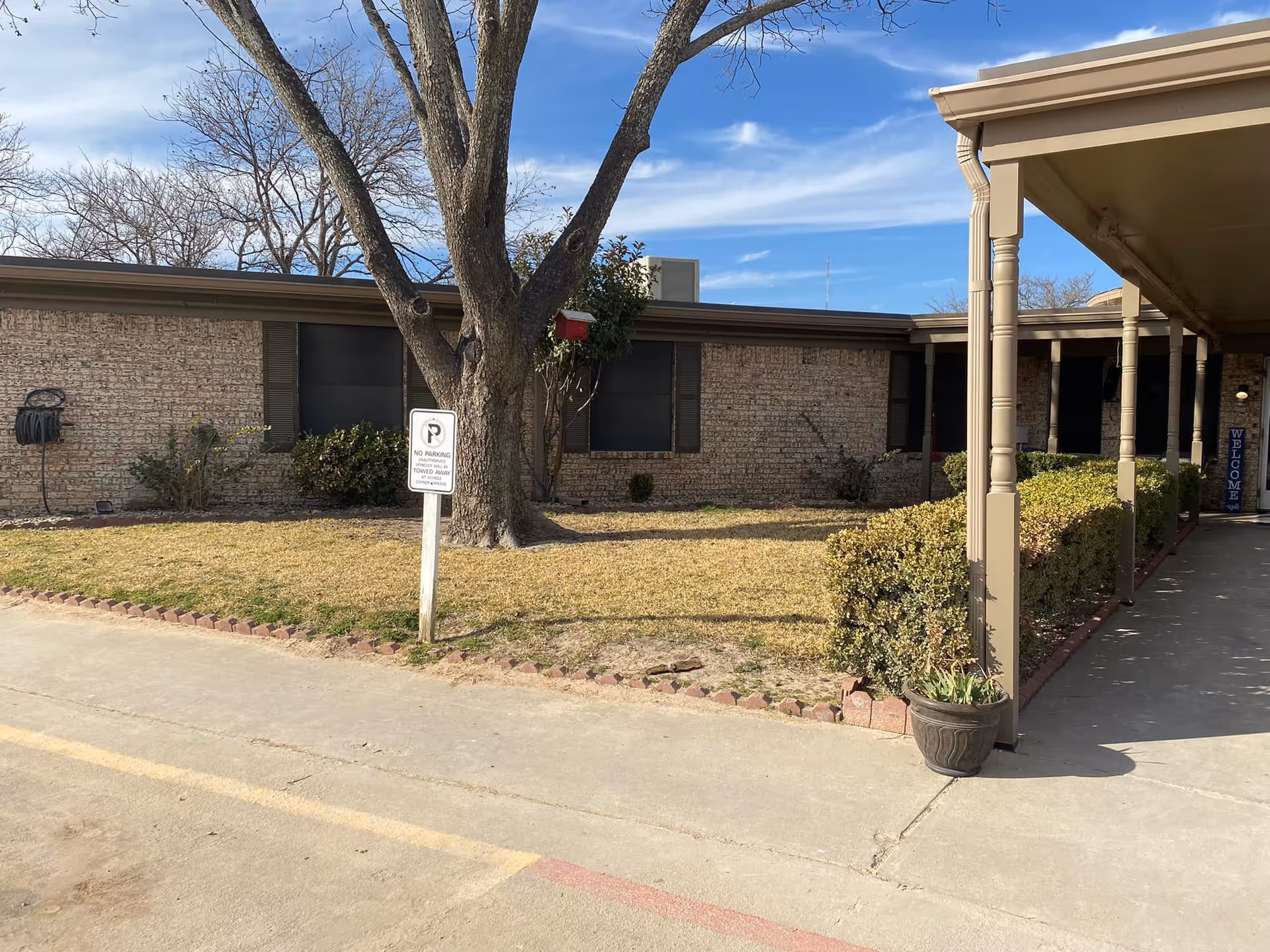 Exterior view of DeLeon Nursing and Rehab facility showing a brick building with several windows, a large tree in a grass area, trimmed bushes along a covered walkway with columns, and a 'No Parking' sign near the tree. The sky is clear with some clouds.