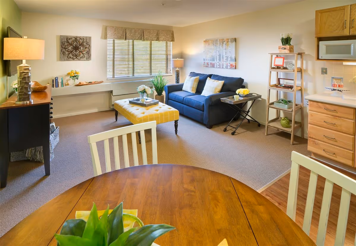 A cozy living area in a senior living facility featuring a blue couch with two pillows, a yellow tufted ottoman, a wooden dining table with white chairs, a small kitchen area with wooden cabinets and a microwave, and decorative plants and artwork on the walls.