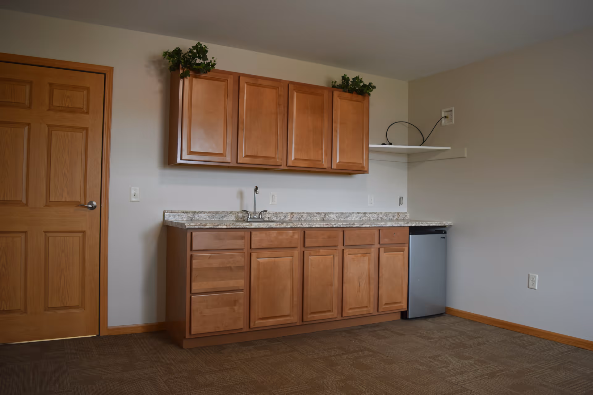 Interior view of a room with wooden cabinets mounted on the wall and below a granite countertop with a small sink. There is a small refrigerator under the counter on the right side and a wooden door on the left. The walls are painted light gray and the floor is carpeted in a brown pattern.