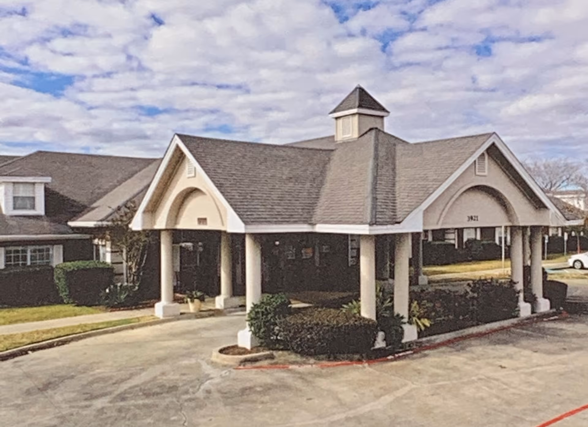 Exterior view of a senior living facility building with a covered entrance supported by columns, surrounded by trimmed bushes and a parking area under a partly cloudy sky.