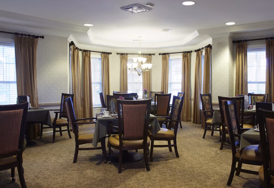 A well-lit dining room with tables set for meals and cushioned wooden chairs arranged near tall windows with gold curtains.