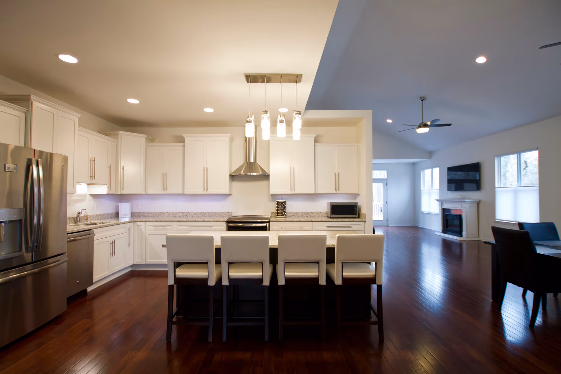 A modern kitchen with white cabinets, stainless steel refrigerator, stove, and dishwasher. There is a kitchen island with four white cushioned chairs. The kitchen opens into a living area with hardwood floors, a ceiling fan, a wall-mounted TV above a fireplace, and large windows letting in natural light.