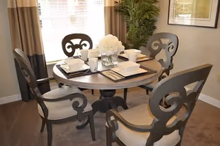 A round wooden dining table set with four ornate chairs in a well-lit room. The table is set with white plates, cups, and napkins, and a vase with white flowers is placed in the center. There is a large window with curtains and a green potted plant in the corner.
