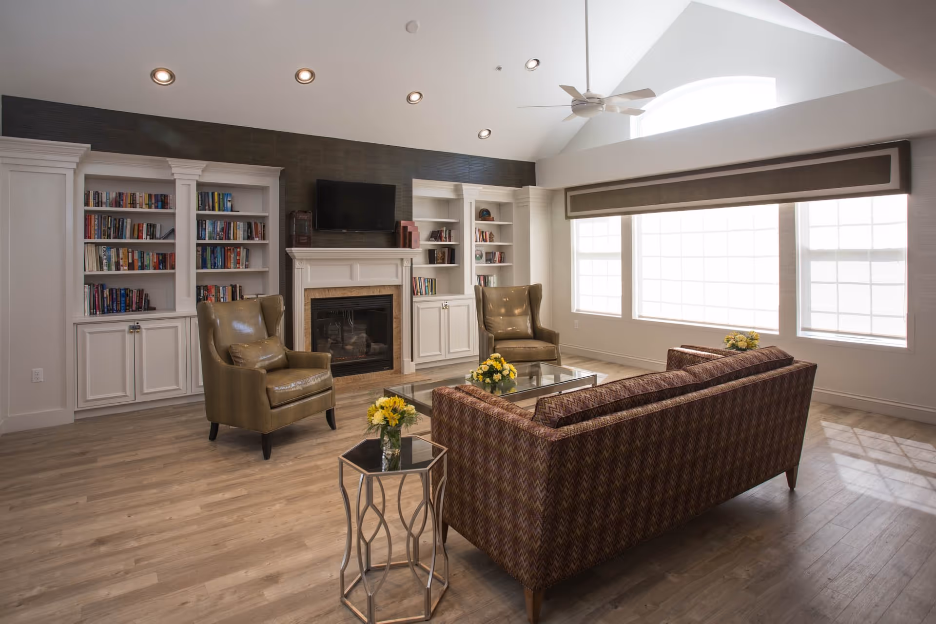 A bright and spacious living room with large windows covered by a valance, allowing natural light to fill the space. The room features a patterned sofa, two leather armchairs, a glass coffee table with a flower arrangement, and built-in white bookshelves flanking a fireplace with a mounted flat-screen TV above it. The ceiling has recessed lighting and a ceiling fan.