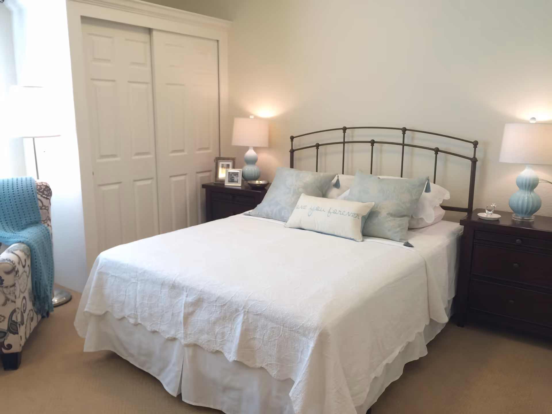 A neatly made bed with a white quilt and three decorative pillows, including one with the words 'love you forever'. The bed has a metal headboard and is flanked by two dark wooden nightstands, each with a blue lamp. To the left, there is a patterned armchair with a blue throw blanket and a floor lamp next to a window. A closed white closet door is visible behind the bed.