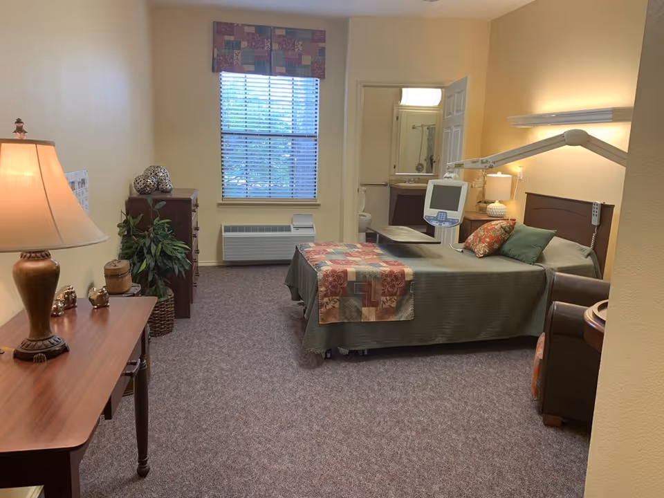 Furnished senior living bedroom with a hospital-style bed and lift, armchair, dresser, lamp, and a window with an en-suite bathroom visible.