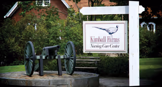 Exterior view of Kimball Farms Nursing Care Center sign with a cannon monument and greenery.