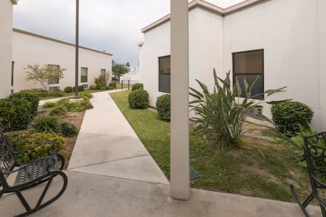 Concrete walkway through a landscaped courtyard between single-story white buildings with benches and shrubs.