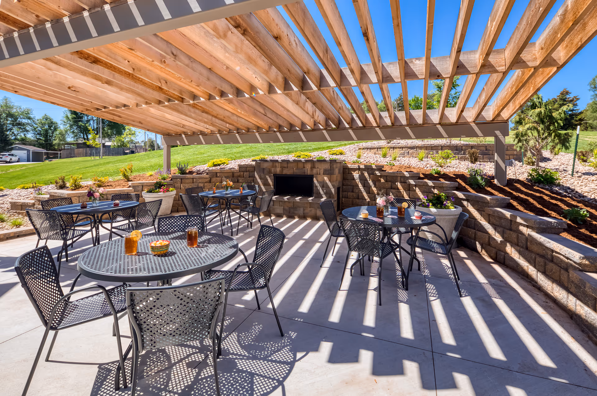 Outdoor patio area with metal tables and chairs under a wooden pergola. Each table has drinks and small bowls or flower arrangements. There is a stone fireplace built into a retaining wall with landscaping and greenery in the background under a clear blue sky.