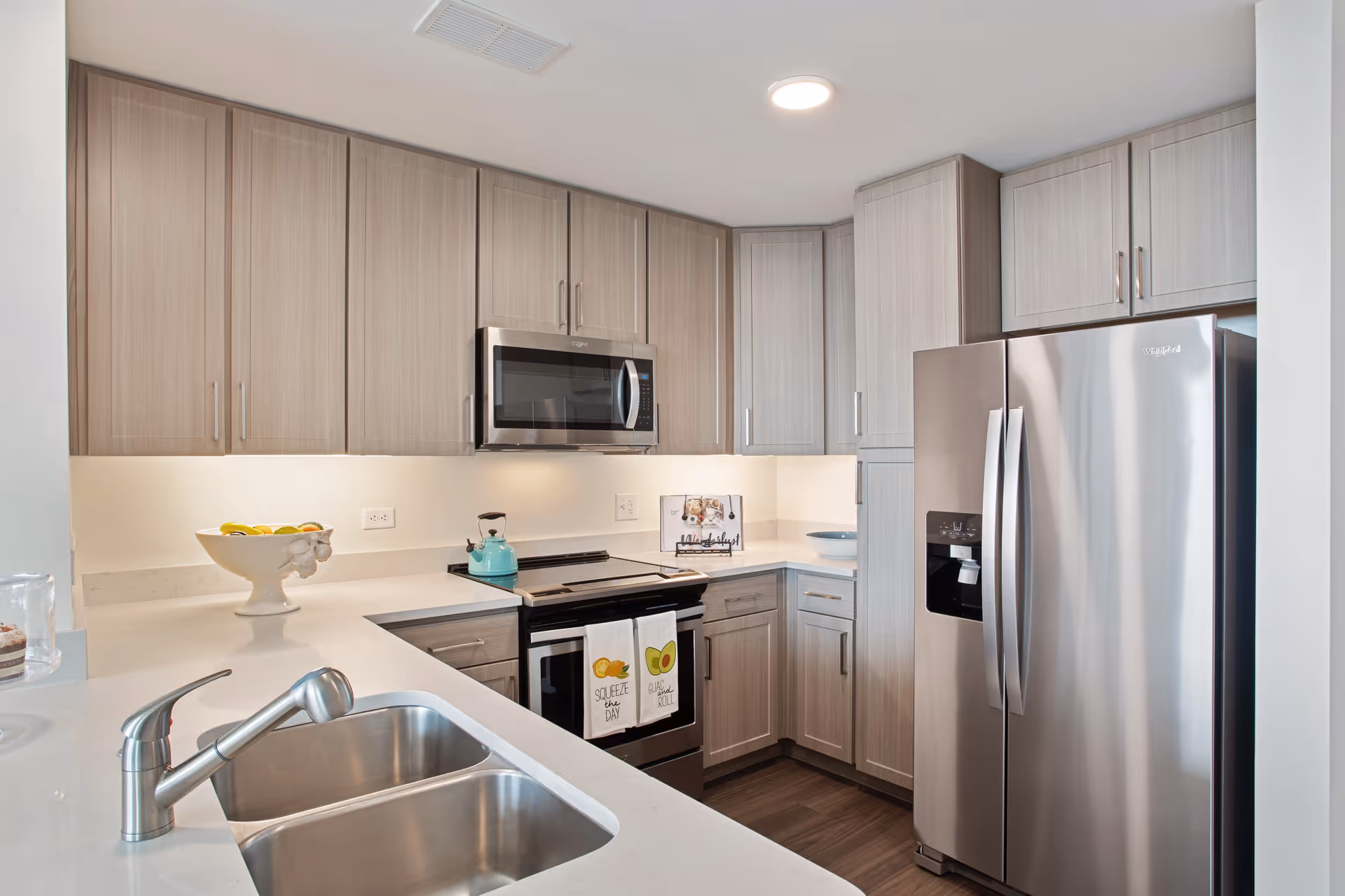 Modern kitchen with light wood cabinets, stainless steel refrigerator, microwave, and stove. White countertops with a double sink and a bowl of fruit on the counter. Two decorative kitchen towels hang on the oven door.