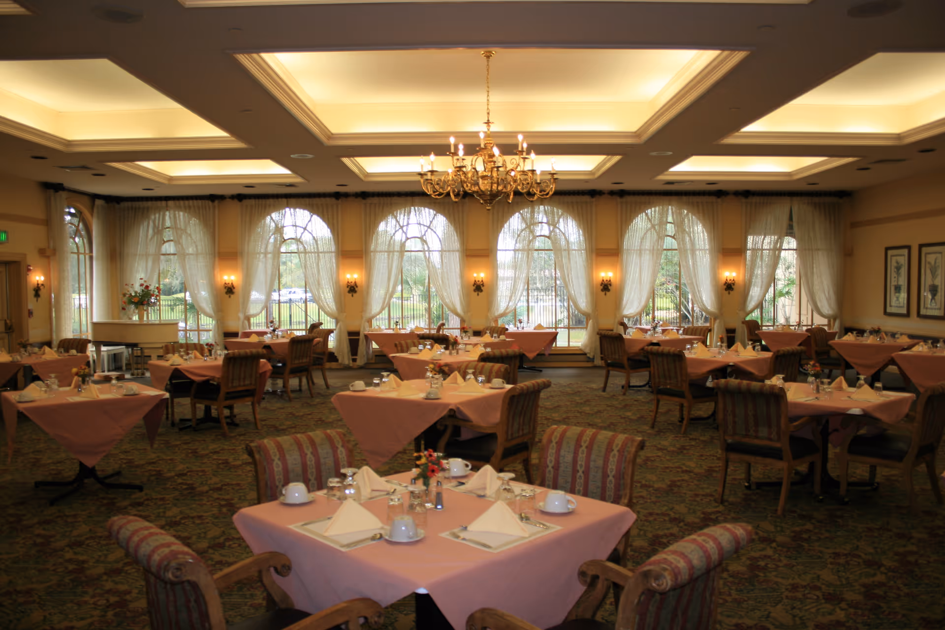 A spacious dining room with multiple tables covered in pink tablecloths, each set with white napkins, cups, and small flower arrangements. The room features large arched windows with sheer curtains, a patterned carpet, and a chandelier hanging from a coffered ceiling.