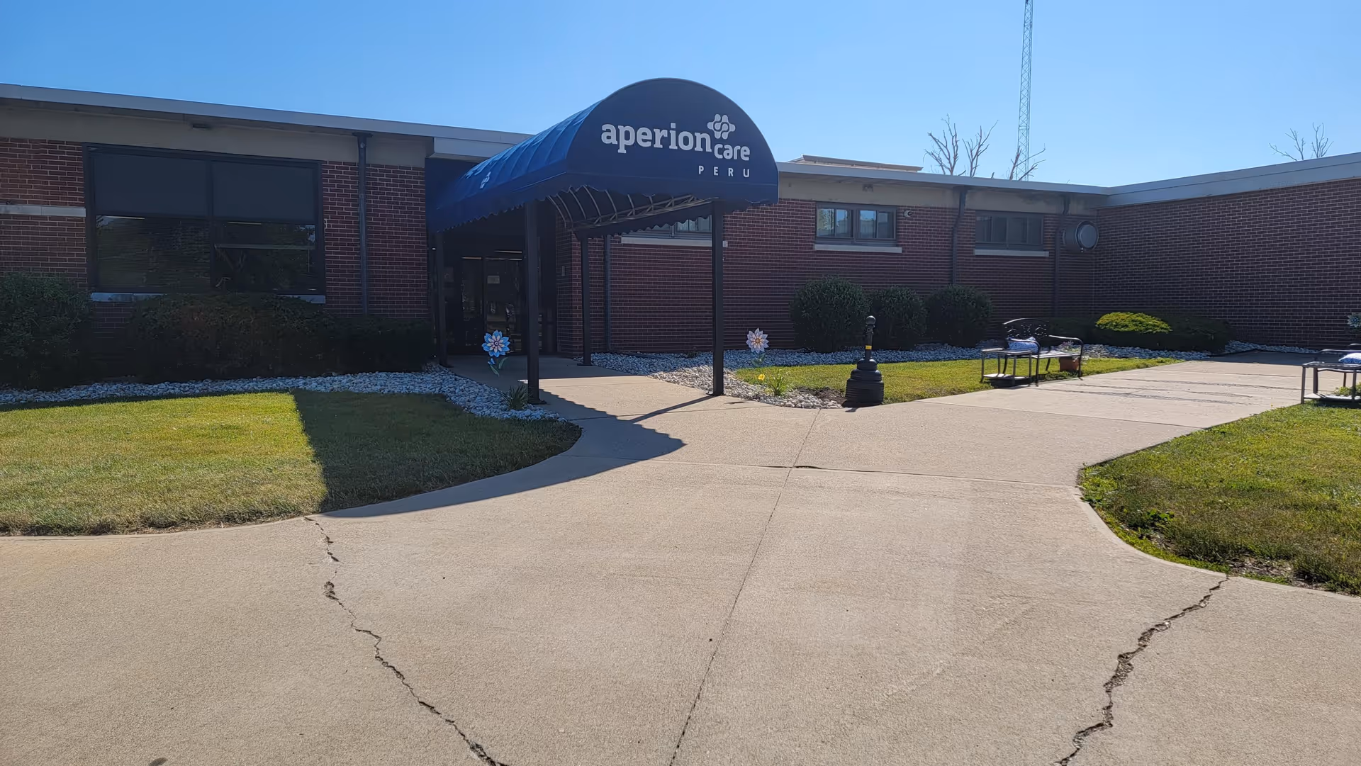 Front entrance of the Aperion Care Peru building with a blue awning over the walkway, surrounding lawn and benches.