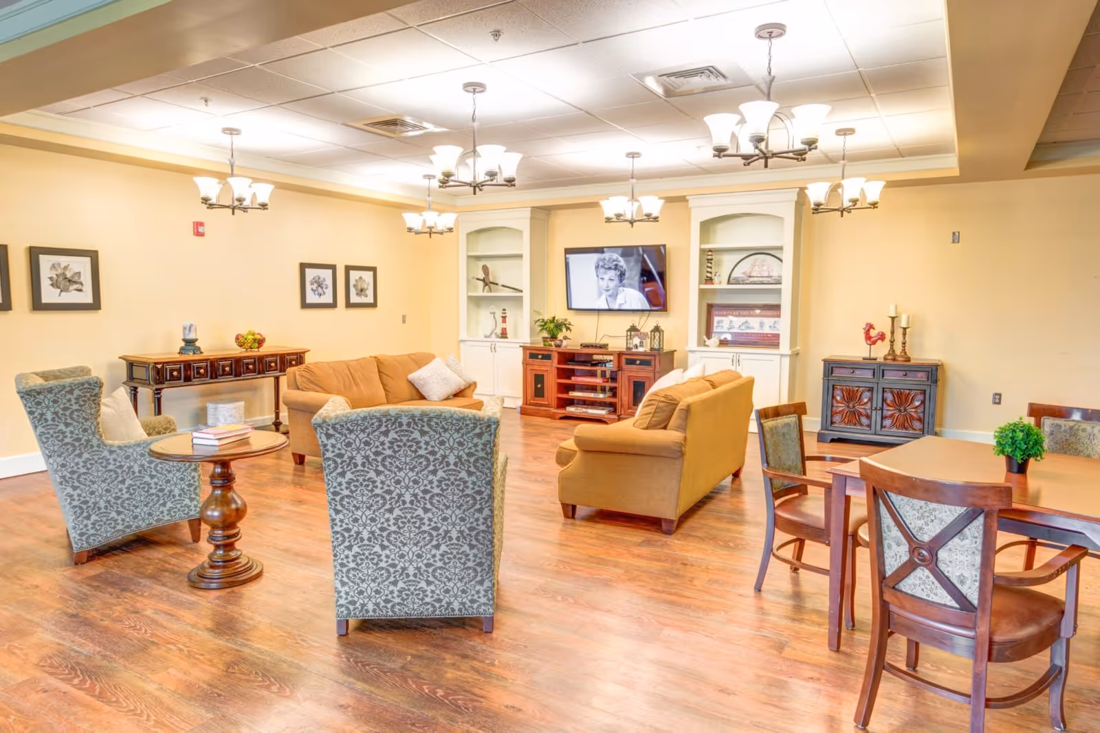 A well-lit living room area with wooden flooring, featuring two beige sofas, two patterned armchairs, a wooden coffee table with books, and a wooden dining table with chairs. The walls are painted light yellow and decorated with framed floral artwork. There are built-in white shelves with decorative items and a flat-screen TV mounted on the wall showing a black and white image of a woman. Multiple ceiling light fixtures provide warm lighting.