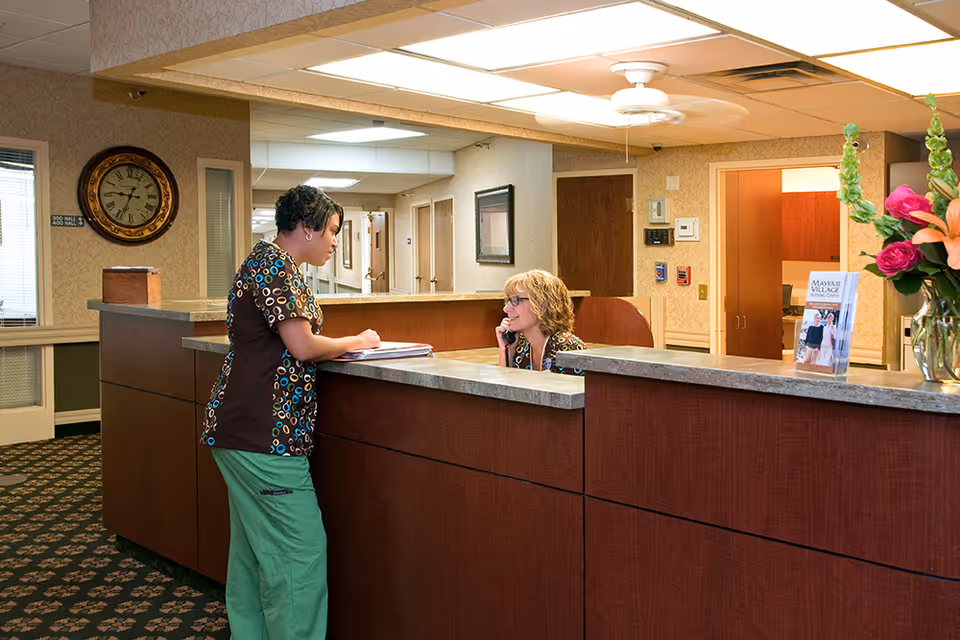 Reception area of Mayfair Village Nursing Care Center with two staff members interacting at the front desk. One staff member is standing and writing on a clipboard, while the other is seated and talking on the phone. The area has a patterned carpet, a wall clock, and a brochure holder with facility information.