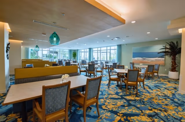 A spacious dining room with multiple tables and chairs arranged neatly. The room features large windows allowing natural light to fill the space, blue and yellow patterned carpet, green pendant lights hanging from the ceiling, and a large painting of a beach scene on the wall. There is also a potted palm tree in the corner.