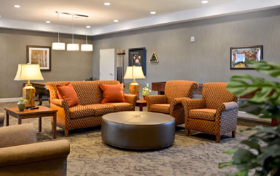 A cozy living room area with a patterned orange sofa and two matching armchairs, a round brown ottoman in the center, side tables with lamps, and framed artwork on the walls. The room has soft lighting and a carpeted floor, with a pool table and cue sticks visible in the background.