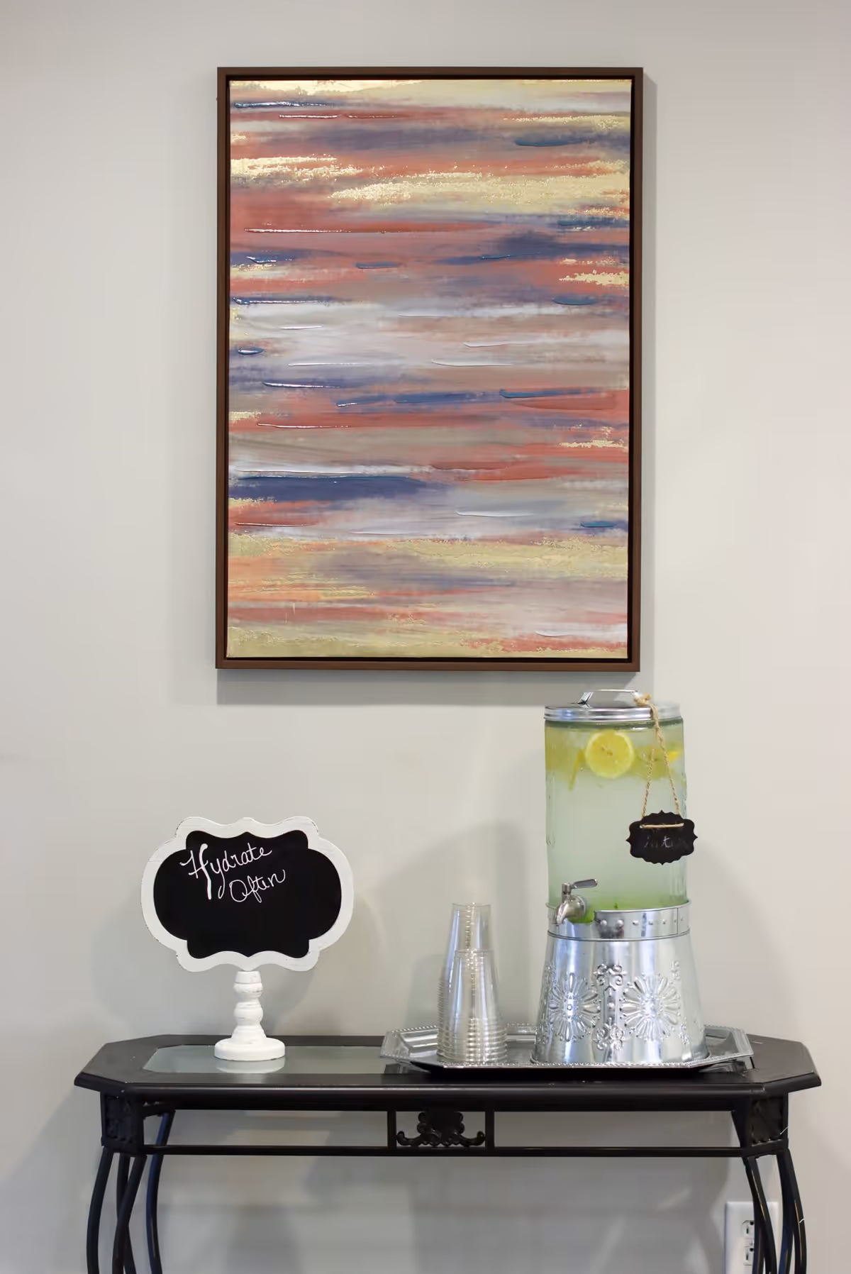 A decorative table against a light-colored wall with a large abstract painting above it. On the table, there is a glass beverage dispenser filled with lemon water, a stack of clear plastic cups on a tray, and a small chalkboard sign that reads 'Hydrate Often.'