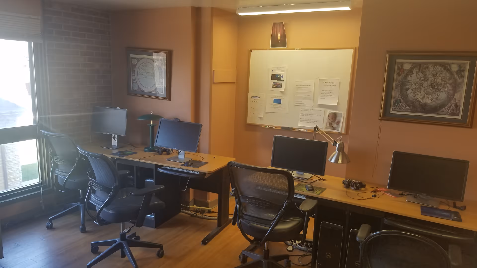 Small computer room with multiple desks, monitors, and office chairs beneath a bulletin board and framed artwork.