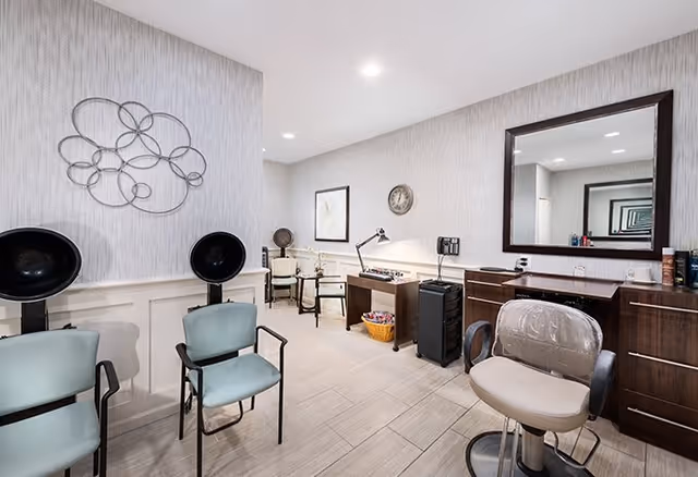 Interior view of a senior living facility hair salon area with two black hair dryers, two green chairs, a styling chair with a protective cover, a large mirror, and a desk with a lamp and hair care products. The room has light-colored tiled flooring and patterned wallpaper with a decorative metal wall art piece.