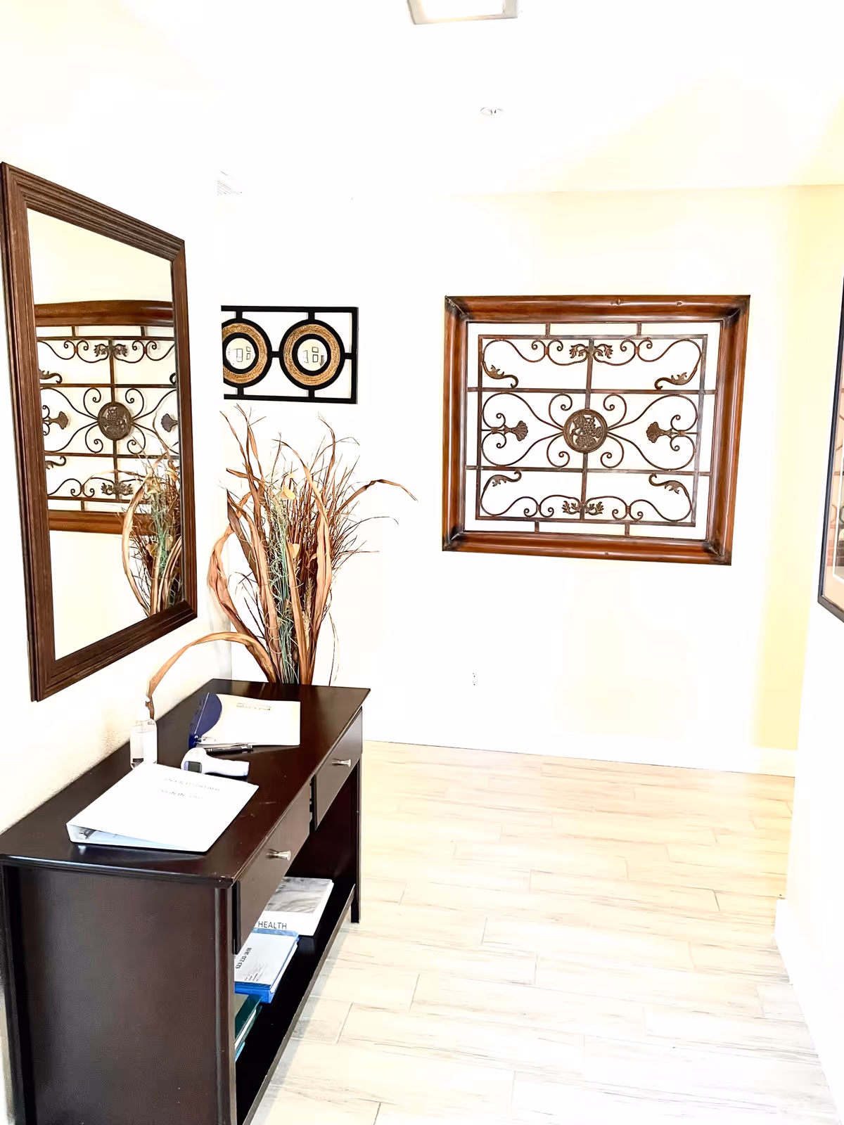 A bright hallway corner with light-colored wooden flooring and white walls. A dark wooden console table with drawers and shelves holds papers, a pen, and a bottle of hand sanitizer. Above the table is a large rectangular mirror with a dark wooden frame. On the wall opposite the mirror is a decorative metal wall art piece framed in dark wood. A tall arrangement of dried plants is placed between the table and the wall art.
