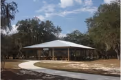 Open-air pavilion with picnic tables under a metal roof and a curved concrete path leading to it amid trees.