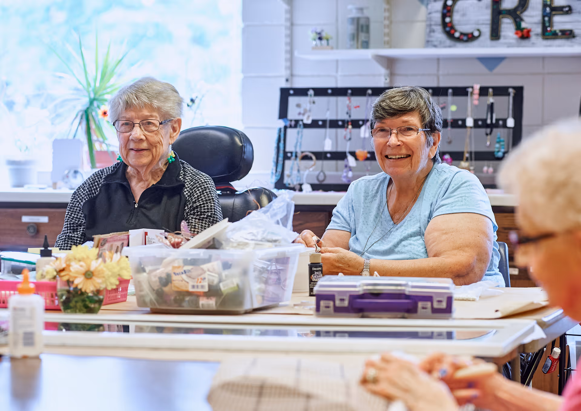 Three elderly women sitting around a table engaged in arts and crafts activities in a well-lit room with shelves and decorations in the background.