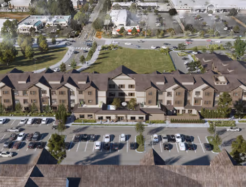 Aerial view of a large senior living facility building with multiple floors and a brown roof. The building is surrounded by parking lots with numerous parked cars, green lawns, and trees. Roads and other buildings are visible in the background.