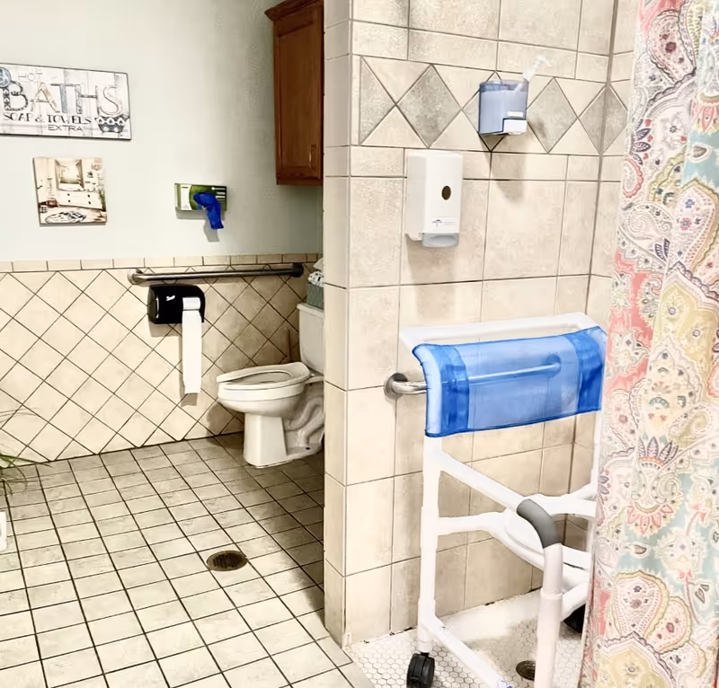 A tiled bathroom with a toilet, a grab bar, a toilet paper dispenser, a blue disposable glove dispenser, and a shower area with a colorful patterned curtain and a white shower chair with blue padding.