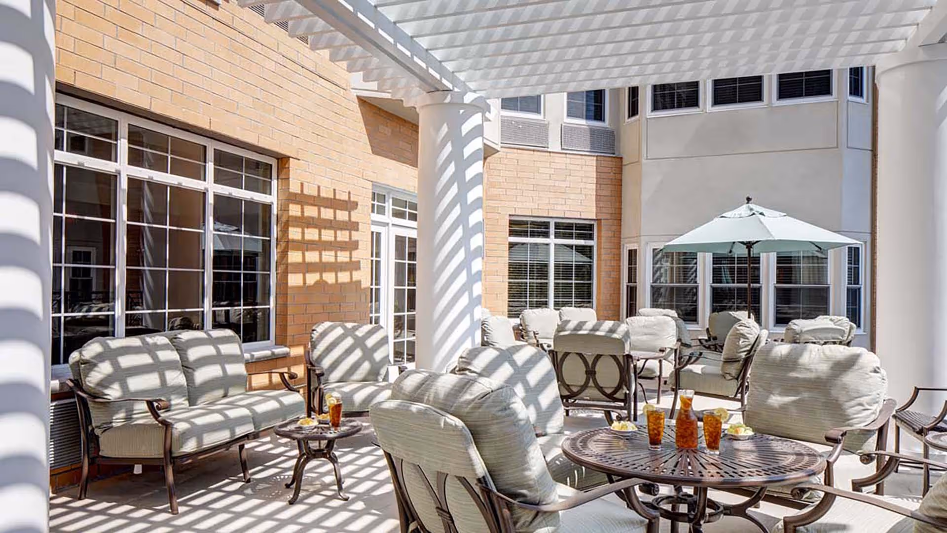 Outdoor patio area with cushioned chairs and tables under a white pergola casting striped shadows. There are drinks and snacks on the tables, and a large umbrella providing shade in the background. The patio is adjacent to a building with large windows and brick and stucco walls.