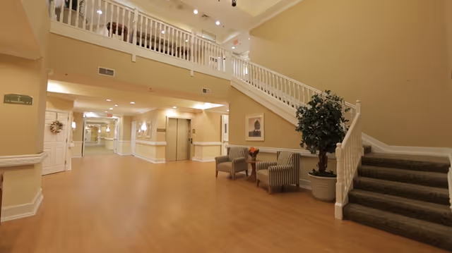 Well-lit senior living lobby with a central elevator, carpeted staircase, seating area, and a potted plant.